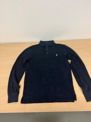 Mörkblå långärmad piké från Polo Ralph Lauren - Säljer en mörkblå långärmad pikétröja från Polo Ralph Lauren med klassisk krage och tre knappar framtill. Tröjan har den ikoniska loggan broderad på bröstet och är i en slim fit-modell. Perfekt för en clean och stilren look.
