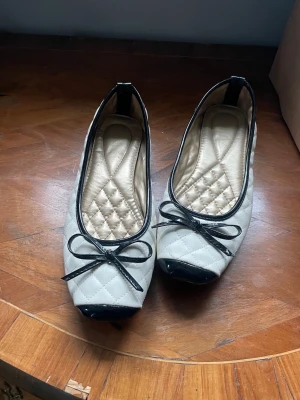 Vita quiltade ballerinaskor med rosett - Snygga ballerinaskor i vitt quiltat material med svarta lackdetaljer på tå och kant. Skorna har en söt svart rosett framtill och en fyrkantig tå. Perfekta lite klassisk och chic 💕💕