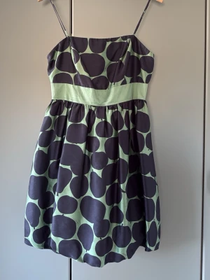 Grön och svart prickig klänning - Snygg kort ballong klänning med tunna axelband och stort prickigt mönster (äpplen och päron) i svart och ljusgrönt. Klänningen har en markerad midja och dragkedja i ryggen. Storlek 6