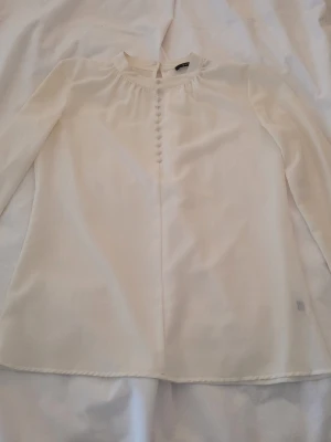 Vit blus med klädda knappar från Esprit - Stilren vit blus från Esprit med rundad halsringning och dekorativa klädda knappar framtill. Blusen har lång ärm och är tillverkad i ett lätt, något transparent tyg som ger ett elegant intryck. Perfekt att matcha med jeans eller kjol.