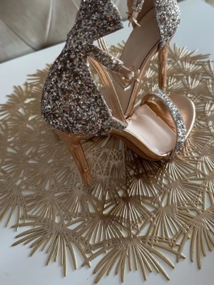 Glittriga guldiga pumps med rem - Säljer ett par glittriga pumps i guld med öppen tå och smal rem över foten. Skorna har höga klackar i metallic och justerbar ankelrem. Perfekta för dig som vill sticka ut med extra bling! Materialet är syntet med glitterdetaljer.