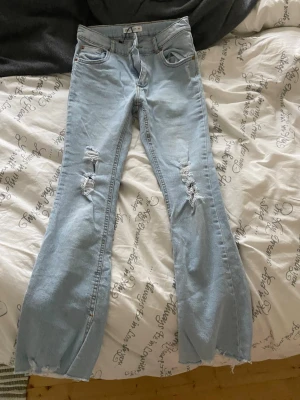 Ljusblå slitna straight/baggy jeans lågmidjade - Säljer ett par ljusblå bootcut jeans med slitningar på låren och trasiga detaljer vid bensluten. Jeansen har klassisk femficksmodell och hög midja. Perfekta för en avslappnad och trendig look.
