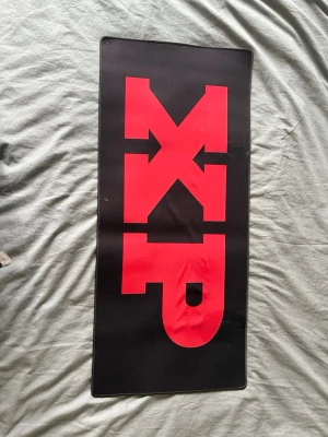 Stor svart och röd XL musmatta - Säljer en stor rektangulär musmatta i svart med stora röda bokstäver 'XP' på ovansidan. Perfekt för gaming eller kontor, ger bra glid för musen och har en halkfri undersida. Tillverkad i textil med gummibotten.