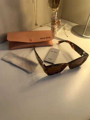 Miu Miu solglasögon brun leopard - Snygga solglasögon från Miu Miu med breda bågar i brun leopardmönstrad plast och stora silvriga logodetaljer på sidorna. Kommer med originalfodral och putsduk. Perfekt accessoar för att lyfta din look.