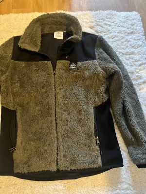 Grå teddyjacka från Warp - Mysig grå teddyjacka från Warp med svarta detaljer på axlar och sidor. Jackan har hel dragkedja, hög krage och två fickor med dragkedja. Perfekt för kalla dagar när du vill vara både varm och stilren.