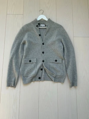 Davida Kashmir Cardigan - Stilren grå kofta från Gant med knappar framtill och två fickor med lock. Koftan har ribbade muddar vid ärmslut och nederkant samt en högre krage. Perfekt för lager på lager och enkel att matcha med olika outfits.