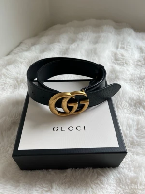 Svart skärp med gulddetalj från Gucci - Snyggt svart skärp från Gucci med den ikoniska dubbla G-spännet i guld. Bältet är gjort i äkta skinn och har en klassisk, stilren design. Spännet är något repigt.
