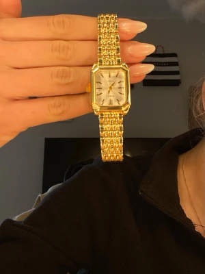 Guldfärgad rektangulär klocka - Stilren guldfärgad klocka med rektangulärt ur och länkarmband. Urtavlan är vit med svarta markeringar och visare. Klassisk design som passar till många olika looks. Metallarmbandet ger en lyxig känsla.