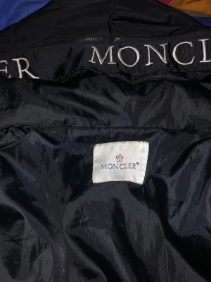 Svart dunjacka från Moncler - Snygg svart dunjacka från Moncler med glansigt material och tydlig logotyp på insidan av kragen. Jackan kan har inga fickor och går inte att dra upp och ner och jackan har ingen sak som man kan dra med upp och ner. Jackan har en klassisk passform och är tillverkad i ett lätt, vindtätt tyg som passar perfekt för blåsiga dagar. 