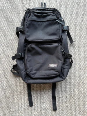 Svart ryggsäck från Eastpak - Säljer en svart ryggsäck från Eastpak med två stora framfickor, justerbara remmar och spännen på sidorna. Tillverkad i slitstarkt tyg och har dragkedjor på alla fack. Perfekt för dig som vill ha en stilren och praktisk väska till skolan eller stan. Ryggsäcken är begagnad och har användningsspår. Släden är skadad på en dragkedja – se bild.