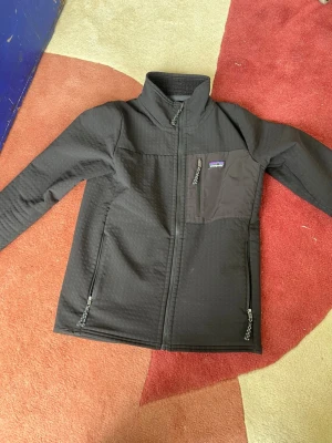 Svart vindjacka från Patagonia - Stilig svart vindjacka från Patagonia med krage, hel dragkedja och tre fickor med dragkedja. Jackan har ett rutmönstrat innerfoder och en bröstficka med logga. Tillverkad i slitstarkt syntetmaterial, perfekt för blåsiga dagar. Var inte rädd för att skriva om intresserad ✅✅