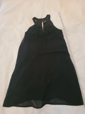 Svart ärmlös kortklänning - Stilren svart kortklänning med halterneck och knapp i nacken. Klänningen är ärmlös och har en lätt A-linjeformad siluett. Tillverkad i ett tunt, flytande material som ger ett elegant fall. Perfekt för dig som gillar enkel och klassisk stil.