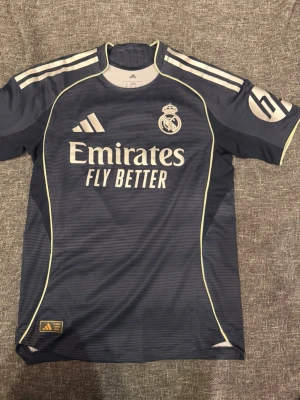 Real Madrid bortatröja 2025/26 authentic - Exklusiv Real Madrids bortaställ 25/26 player edition med Valverde tryck på ryggen. I super bra skick som ny då den använts enstaka gång. Nypris 1600kr, säljer för 800kr då den är som ny. Adidas Real Madrid fotbollströja i mörkblå färg med ljusa detaljer. Tröjan har klubbmärke på bröstet, Adidas-logga och sponsortryck på framsidan. På ryggen finns namntryck "Valverde" och nummer 8. Storlek S enligt märkning. Kortärmad modell i funktionsmaterial som passar för fotboll och träning eller vardagligt