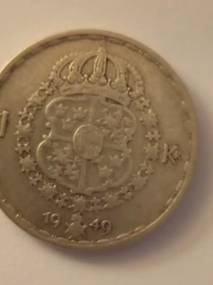 Svensk enkrona 1949 Gustaf V - Säljer en svensk enkrona från 1949 med Gustaf V:s porträtt på ena sidan och Sveriges riksvapen med krona på andra sidan. Myntet är i silverfärgat metall och har texten 'GUSTAF V SVERIGES KONUNG' samt 'MED FOLKET FÖR FOSTERLANDET' runt kanten.
