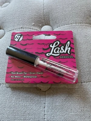 W7 Lash Adhesive ögonfranslim - W7 Lash Adhesive är ett genomskinligt ögonfranslim med tunn borste som gör appliceringen enkel och exakt. Limmet är vattenfast och torkar klart, perfekt för lösögonfransar. Förpackningen är rosa med svarta detaljer och innehåller 5 ml. Oanvänd 