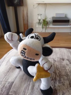 Mjukistjur med stjärna - Rolig mjukistjur i svart och vitt med broderat ansikte och ett stort leende. Tjuren har svarta horn, svans och öron samt håller i en gul filtstjärna. Tillverkad i mjukt tyg, perfekt som dekoration eller leksak.