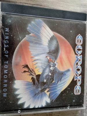 Europe Wings of Tomorrow CD - CD-skiva med albumet 'Wings of Tomorrow' av Europe. Omslaget har en illustration av en örn mot en röd planet och rymdbakgrund. Skivan är silverfärgad med svart text och Epic-logga. Innehåller låtar som 'Dreamer' och 'Dance the Night Away'.