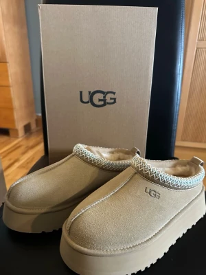 Beige UGG tofflor med platåsula - Säljer ett par beige UGG tofflor med platåsula och mjukt foder. Tofflorna har en broderad kant runt öppningen och UGG-loggan på sidan. Tillverkade i mocka med en chunky sula som ger extra höjd och en trendig look. Helt nya! Skorna har äktighetsintyg med qr kod. 