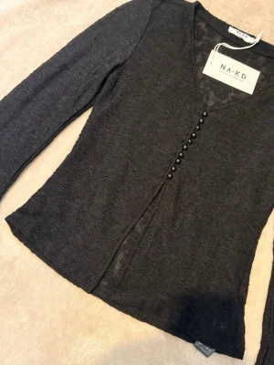 Svart tunn kofta från NA-KD - Snygg svart tunn kofta från NA-KD med v-ringning och dekorativa knappar framtill. Modellen är figurnära med långa ärmar och lätt transparent material som ger en trendig look. Perfekt att styla över ett linne eller topp.