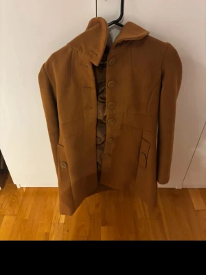Brun ullrock med stora knappar - Snygg brun rock i ull med stora dekorativa knappar framtill och klassisk krage. Rocken har långa ärmar, två fickor med lock och ett slätt innerfoder. Perfekt för kyligare dagar och ger en stilren look.
