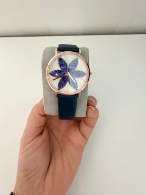 Blå klocka från Ally Denovo - Stilren klocka från Ally Denovo med blått armband och urtavla dekorerad med ett stort blommotiv i blått och vitt. Boetten är i guldton och armbandet är i ett mjukt material. Perfekt accessoar för att lyfta din outfit.