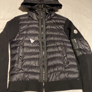 Svart pufferjacka från Moncler - Snygg svart pufferjacka från Moncler med stickade ärmar och rygg. Jackan har huva, två sidofickor med dragkedja och en Moncler-logga på vänster ärm. Framsidan är quiltad och materialet är en mix av dun och stickat tyg. Storlek M men sitter litet så passar jättebra som S