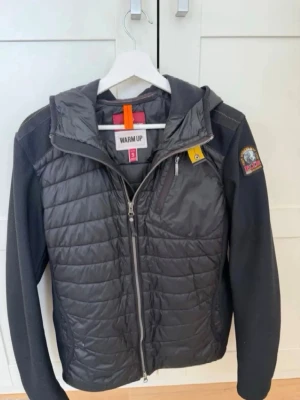 Svart pufferjacka från Parajumpers - Snygg svart pufferjacka från Parajumpers med quiltad front och rygg, mjuka stickade ärmar och huva. Jackan har dragkedja framtill, bröstficka med zip och ikonisk gul detalj vid halsen. Perfekt för kalla dagar och riktigt stilren.