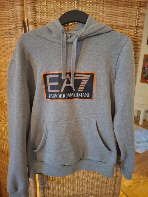 Grå hoodie från Emporio Armani EA7 - Snygg grå hoodie från Emporio Armani EA7 med stor logga på bröstet i svart, vitt och orange. Klassisk känguruficka framtill, justerbar huva med dragsko och ribbade muddar. Perfekt för en avslappnad och trendig look.