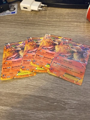 Fyra Charizard Pokémonkort - Säljer fyra Charizard Pokémonkort med eldig design och holografisk effekt. Korten har starka attacker som Flame Cloak och Burning Breath. Perfekt för samlare eller dig som vill förstärka din kortlek. Alla kort är officiella och har klassisk Pokémon-baksida.