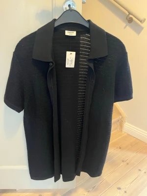 Svart stickad kortärmad skjorta Kappahl - Svart stickad skjorta från Kappahl med korta ärmar och klassisk krage. Koftan har knappar framtill och är tillverkad i ett luftigt, lätt material som passar perfekt till lager på lager