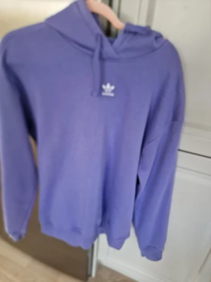 Lila hoodie från Adidas - Säljer en lila hoodie från Adidas med den klassiska loggan broderad i vitt på bröstet. Hoodien har huva med dragsko och ribbade muddar vid ärmar och nederkant. I mycket gott skick. Lappen är borta.  Storlek S eller M