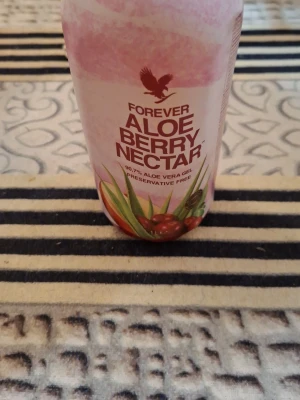 Forever Aloe Berry Nectar dryck - Säljer en flaska Forever Aloe Berry Nectar, en dryck med 90,7% aloe vera gel och bärsmak. Flaskan är 1 liter och har en rosa och röd design med illustrationer av aloe vera och bär. Perfekt för dig som gillar hälsodrycker utan konserveringsmedel.