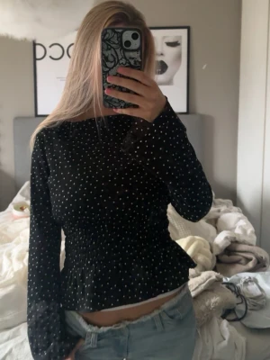Svart prickig blus med volang - Säljer en svart blus med vita prickar och smockad midja som ger en snygg form. Blusen har långa ärmar och volang nertill för en extra touch. Perfekt till jeans eller kjol för en trendig look.