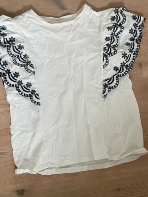 Vit blus med broderade volangärmar - Jättesnygg vit volangblus från zara med svart broderi. Storleken är 164 vilket anses passa för 13-14 åringar. Blusen har ett skönt tyg som gör den väldigt skön att ha på sig. Perfekt för somriga dagar men även till vardags och finare tillfällen.  Tröjan har inga defekter eller fläckar.