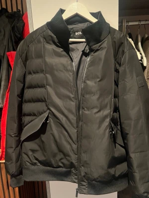 Svart pufferjacka från Hugo BOSS - Snygg svart pufferjacka från BOSS med diskret logga på ärmen. Jackan har hög krage, dragkedja framtill och två stora fickor med lock. Materialet är glansigt och quiltat för extra värme och stil. Perfekt för kalla vinterdagar och ger en clean look. Använd max 1-2 gånger. Köpt för betydligt dyrare. Slutsåld hos Hugo Boss