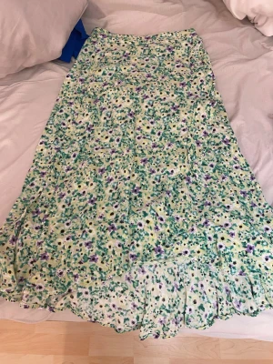 Blommig långkjol med volangkant - Säljer en långkjol med blommigt mönster i grönt, lila och vitt. Kjolen har en volangkant nedtill och är gjord i ett lätt, flowy tyg som känns perfekt för vår och sommar. Midjemodellen är elastisk för extra komfort. Det är i skorlek 128 men passar för mig som är 34,xs/s i byxor