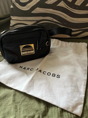 Svart Marc Jacobs crossbody en special edition  - Snygg svart handväska från Marc Jacobs med guldfärgad logga framtill. Väskan har en kompakt levereras med original dustbag. Perfekt för dig som gillar stilrena accessoarer.