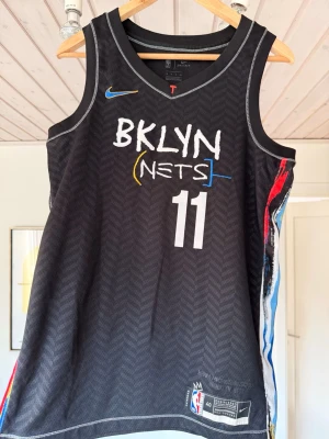 Brooklyn Nets basketlinne Irving #11 - Snyggt svart basketlinne från Nike med Brooklyn Nets tryck och nummer 11, samt Irving på ryggen. Linnet har färgglada detaljer i blått, rött och gult på sidorna och är tillverkat i ett lätt och ventilerande material.  köpt från nike direkt