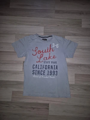 Ljusblå t-shirt med tryck South Lake - Snygg ljusblå t-shirt med trycket 'South Lake State Park California Since 1993' i rött, vitt och svart framtill. Klassisk passform och rund halsringning. Perfekt för en avslappnad stil. Den har 2 små hål fram,  se bild.