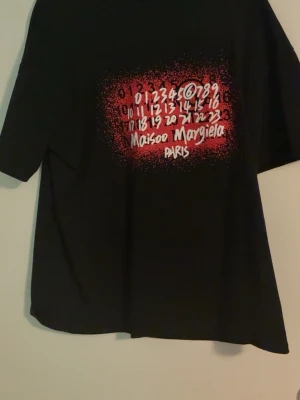 Maison Margiela svart t-shirt - Cool svart t-shirt från Maison Margiela med grafiskt tryck i vitt och rött framtill. Trycket har siffror och Maison Margiela Paris-loggan i street art-stil. T-shirten har rund hals och korta ärmar, perfekt för en avslappnad och trendig look.