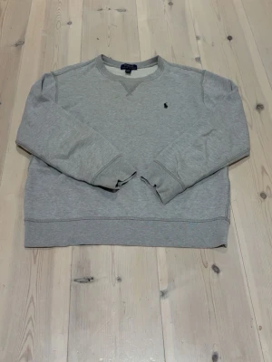 Grå sweatshirt från Polo Ralph Lauren - Klassisk grå sweatshirt från Polo Ralph Lauren med rund halsringning och diskret broderad logga på bröstet. Tröjan har ribbade muddar vid ärmslut och nederkant samt en relaxed passform. Perfekt för en avslappnad och stilren look.