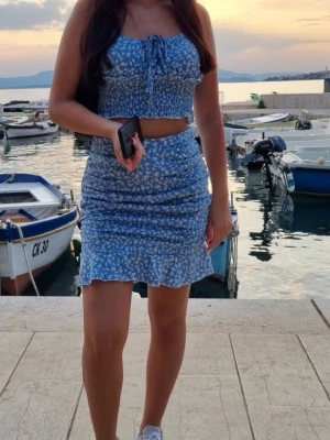 Blått blommigt set - Blått set med blå croppad linnetopp med små vita blommor och knyt framtill. Toppen har smala axelband och en figurnära passform. Matchande kjol som också är figurnära.