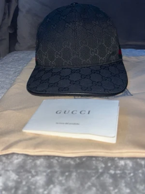 Svart keps från Gucci med GG-mönster - Säljer en svart keps från Gucci med det klassiska GG-monogrammönstret över hela kepsen. Kepsen har böjd skärm, justerbart spänne bak och diskreta röda och gröna detaljer på sidorna. Tillverkad i canvasmaterial med exklusiv känsla.