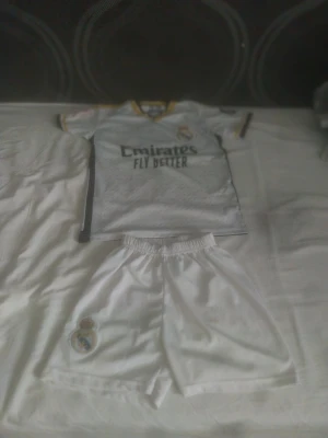 Real Madrid matchställ vit/gul - Säljer ett Real Madrid matchställ med vit tröja och shorts. Tröjan har svarta och gula detaljer, klubbmärke på bröstet och sponsortryck framtill. Shortsen är vita med klubbmärke på benet. Perfekt för fotbollsträning eller match.