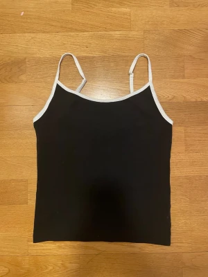 Svart linne med vita kanter BikBok - Säljer ett svart linne från BikBok med tunna vita axelband och vita kanter. Enkel och stilren design, perfekt för lager på lager eller till jeans. Materialet är mjukt och stretchigt för en bekväm passform.