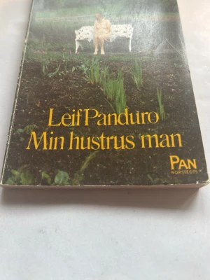 Min hustrus man av Leif Panduro - Romanen 'Min hustrus man' av Leif Panduro är en dansk skönlitterär bok i pocketformat. Omslaget är färgstarkt med en bild av en man på en bänk i en trädgård. Boken är tryckt av PAN/Norstedts och har ISBN 91-1-741251-x.