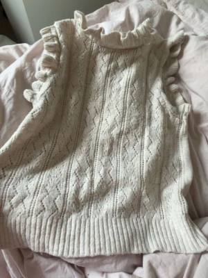 Vit stickad ärmlös topp med volang - Säljer en söt vit stickad topp med volangdetaljer vid axlarna och halsen. Toppen är ärmlös och har ett diskret hålmönster framtill som ger en trendig look. Perfekt att styla med jeans eller kjol för en fräsch outfit.