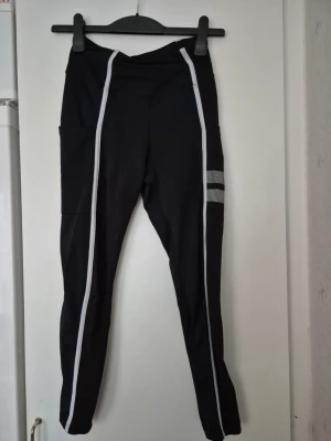 Träningstights - Svarta träningstights från Stronger med vita vertikala ränder längs benen och reflekterande detaljer på ena benet. Tightsen har hög midja, stretchigt funktionsmaterial och praktiska sidofickor. Perfekta för gym, löpning eller annan sport.