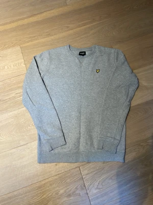 Grå sweatshirt från Lyle & Scott - Klassisk grå sweatshirt från Lyle & Scott med rund halsringning och deras ikoniska gula örnlogga broderad på bröstet. Tröjan har ribbade muddar vid ärmslut och nederkant. Perfekt för en avslappnad och stilren look. Junior storlek typ som ett S