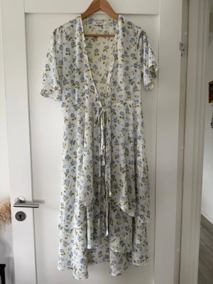 Vit blommig omlottklänning NA-KD - Lång, vit omlottklänning med blått och gult blommönster. Klänningen har korta ärmar, v-ringning och knytband i midjan. Tillverkad i ett lätt och luftigt material, perfekt för varma dagar. Midi klänning 
